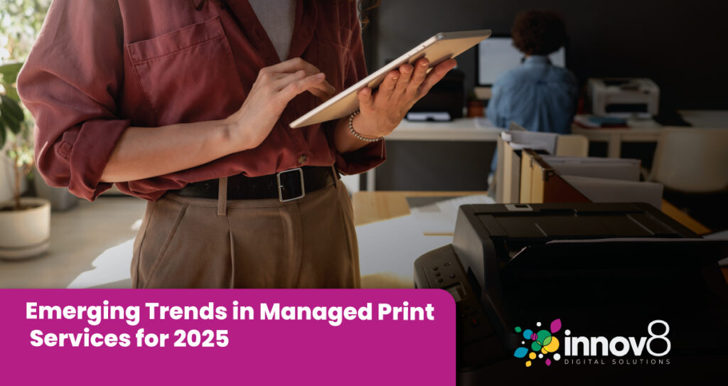 A professional woman wearing a maroon shirt and khaki pants holding a tablet, standing near a printer in a modern office environment.