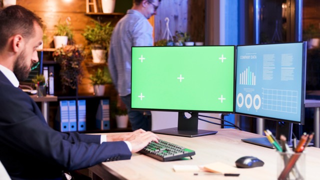 A businessman in a formal suit working on a desktop computer with dual monitors, one displaying a green screen and the other showing business data, in a modern office setting.