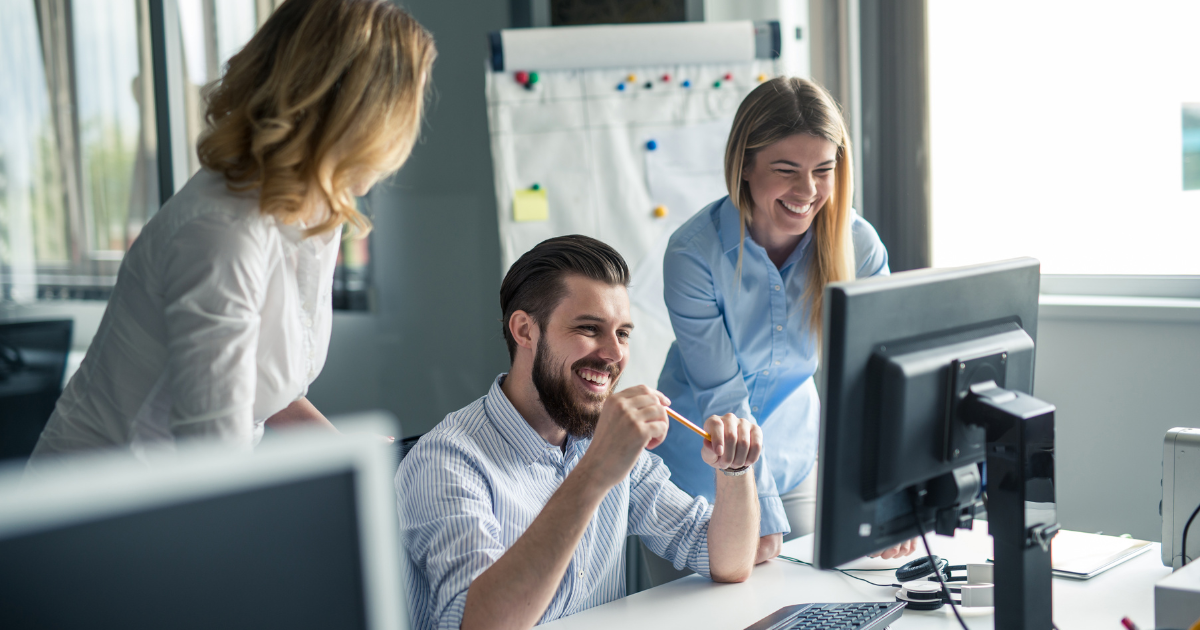 Team of professionals smiling and collaborating around a desktop computer in a bright modern office.