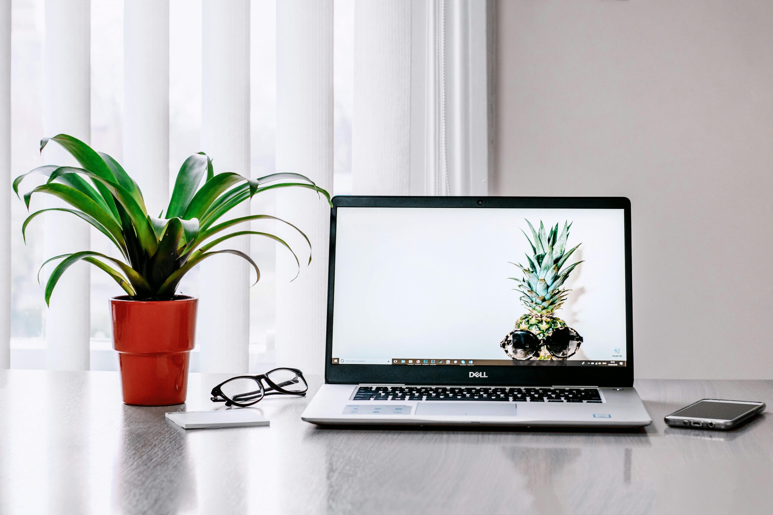 Open laptop on a desk with a pineapple wallpaper, next to a potted plant, glasses, notebook, and smartphone.