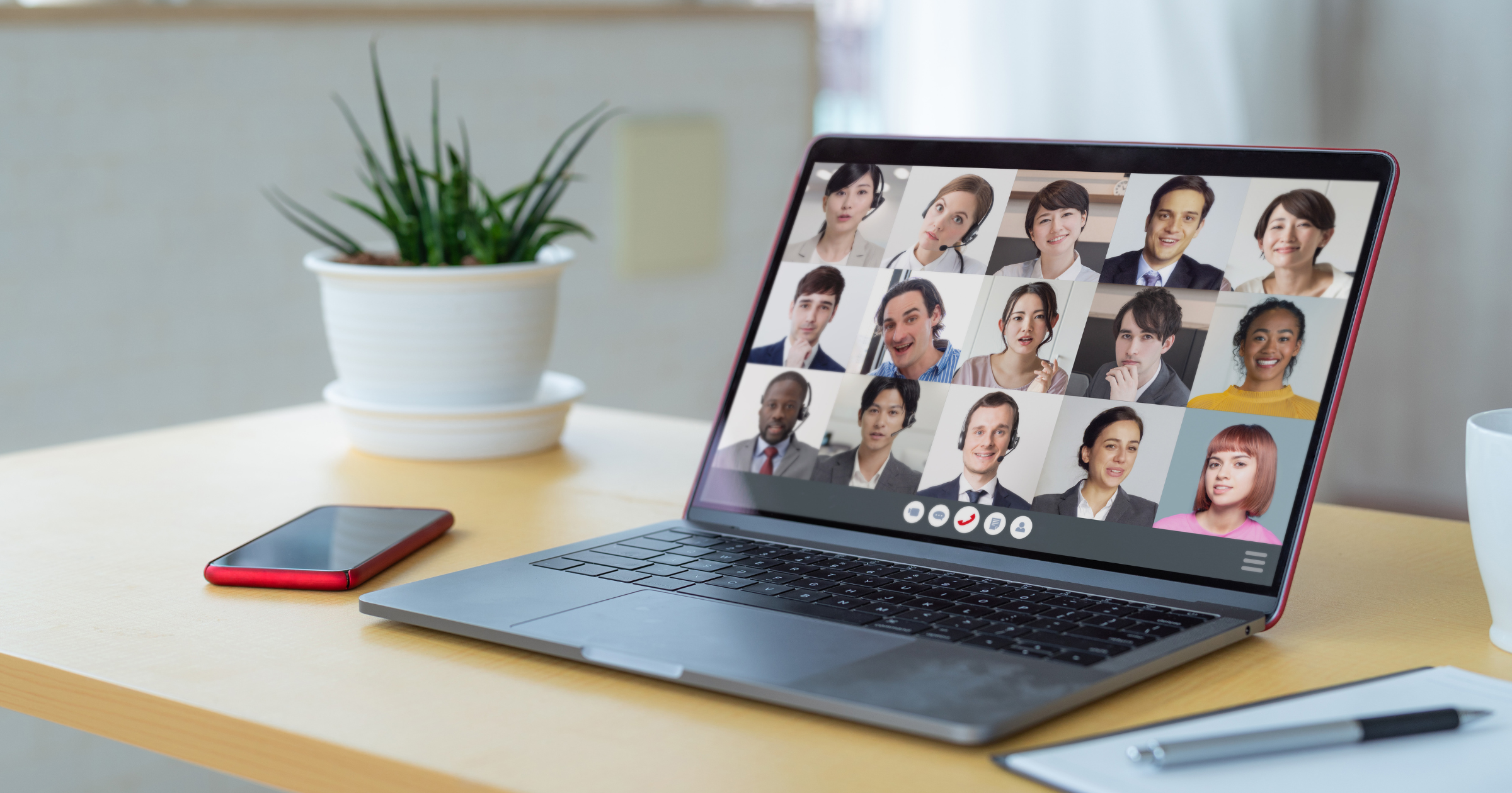 Laptop on a wooden desk displaying a video conference with diverse participants, alongside a plant, smartphone, and coffee mug.