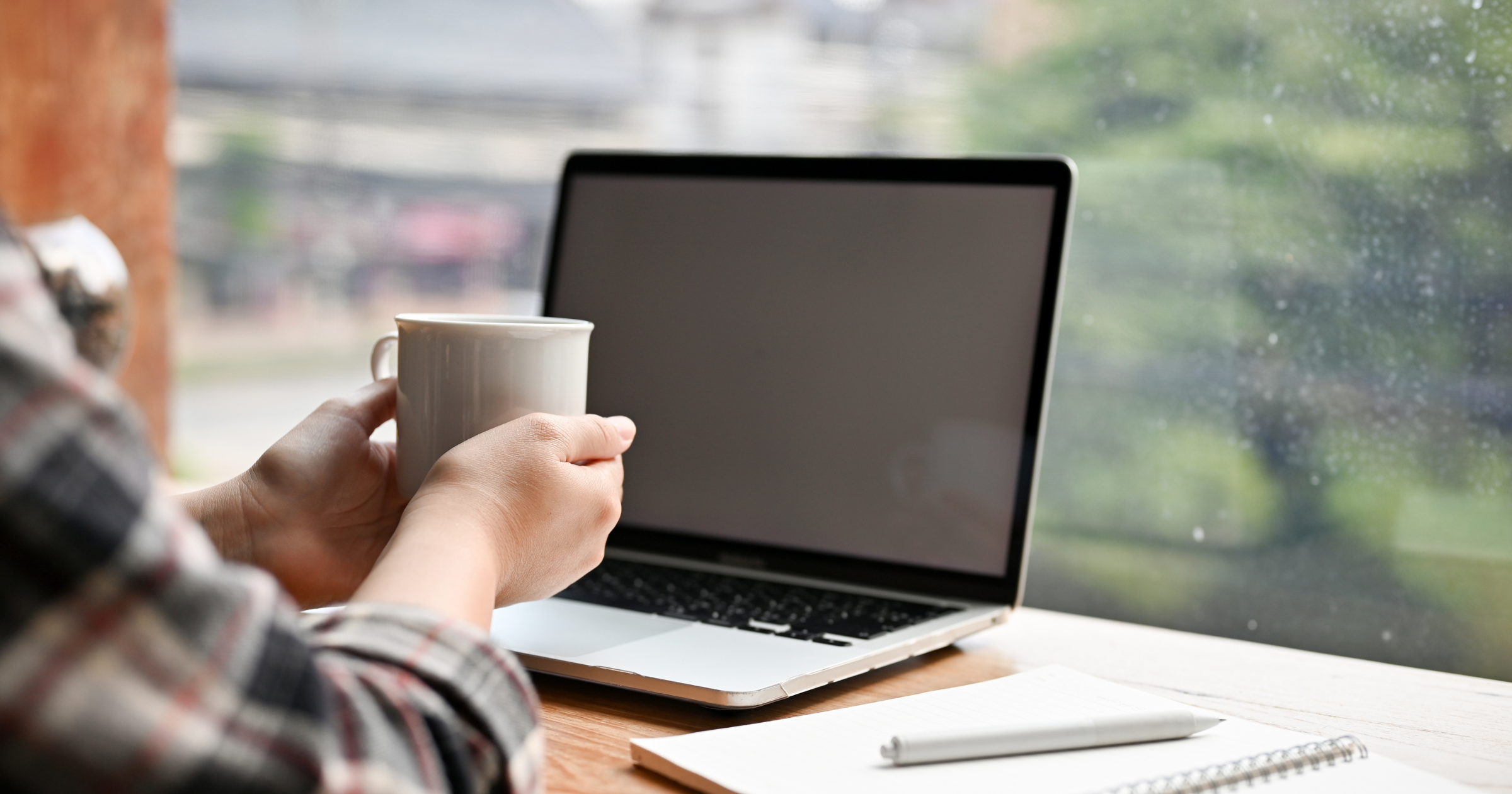A person holding a white coffee mug in front of a closed laptop on a wooden desk, with a notebook and pen nearby, next to a window with a blurred outdoor view.