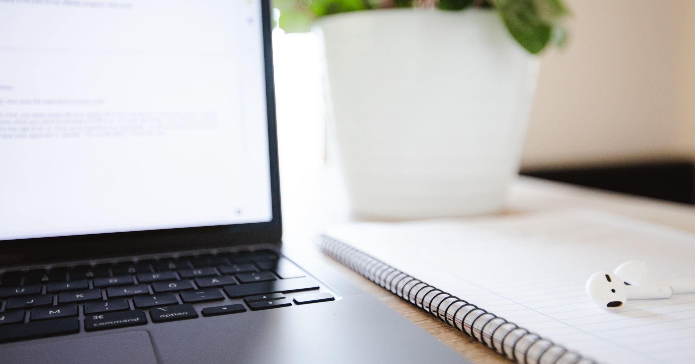 Close-up of a remote work setup with a laptop keyboard, a notebook, wireless earphones, and a white potted plant on a desk.