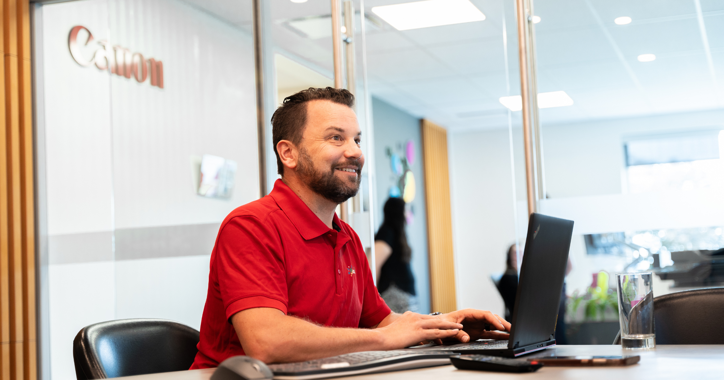 Smiling man in a red polo shirt working on a laptop in a modern office setting with glass walls and a Canon logo in the background.